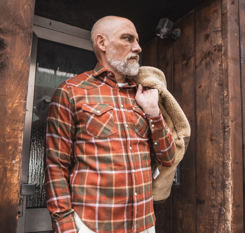 Uomo con camicia western in flanella a quadri e giacca con collo in montone sulla spalla, davanti a un portone di legno. Foto @chesapeakesofficial