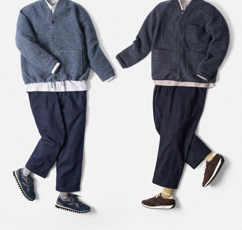 Due completi Universal Works stesi in piano: cardigan in lana blu scuro e blu chiaro, camicia bianca, pantalone blu e sneakers.